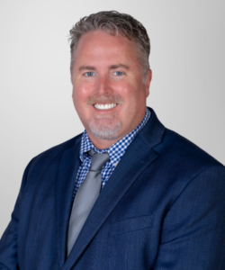 A man with short, light brown hair and a beard is smiling, dressed in a blue suit with a gray tie and blue checkered shirt, standing against a light gray background, representing professionalism in revenue cycle management.