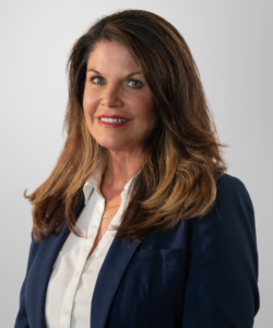A woman with long brown hair, wearing a navy blazer over a white blouse, smiles confidently in front of a plain light gray background, reflecting professionalism often associated with revenue cycle management.