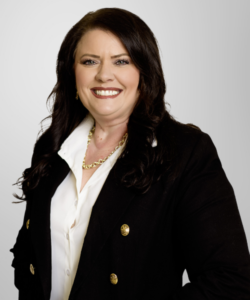 A woman with long dark hair wearing a black blazer, white blouse, and gold necklace smiles at the camera against a plain light background, embodying professionalism in revenue cycle management.