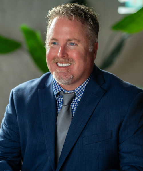 Brandon Walters, a smiling man with short light hair and a trimmed beard, wears a blue suit, checkered shirt, and gray tie. He is seated indoors with blurred green plants in the background.