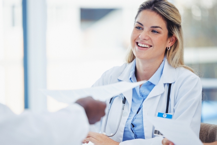 A smiling female doctor in a white coat and stethoscope sits at a desk, handing or receiving chargemaster paperwork from another person in a brightly lit office.