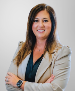 A woman with long brown hair in a beige blazer and black top, smiling with arms crossed, stands against a light gray background. She wears a smartwatch and delicate necklace—confidently representing revenue cycle management expertise.