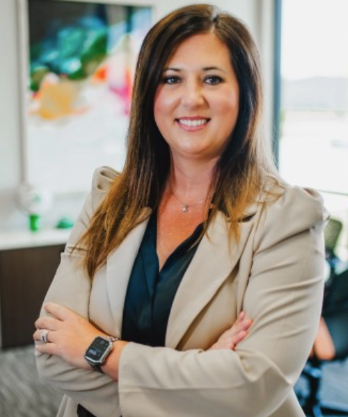 A woman with long brown hair in a beige blazer and black top, smiling with arms crossed, stands against a light gray background. She wears a smartwatch and delicate necklace—confidently representing revenue cycle management expertise.