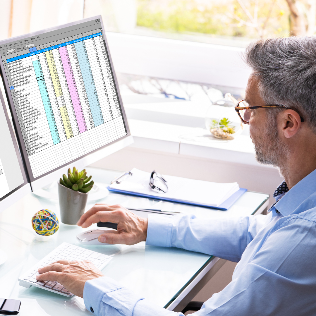 A man with gray hair and glasses works at a computer displaying a colorful spreadsheet, exploring Our Services. He is seated at a glass desk with office supplies, a notebook, and small plants nearby.