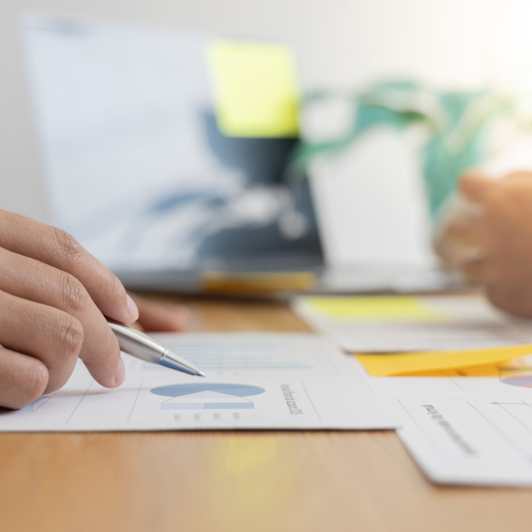 Close-up of a person holding a pen and pointing at a printed chart on a desk, with documents, a laptop, and another person’s hand visible in the background—illustrating how Our Services support informed decision-making.