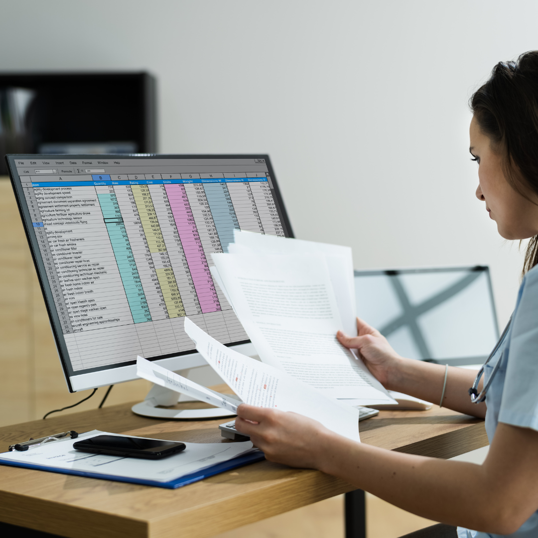 A woman sits at a desk reviewing documents while looking at a computer screen displaying a colorful spreadsheet, focused on managing Our Services. She is surrounded by papers, a clipboard, and a smartphone.