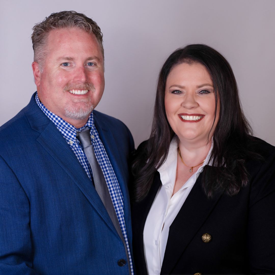 A man in a blue suit and tie stands next to a woman in a dark blazer and white blouse. Both are smiling and posing in front of a plain light background, capturing the beginning of Our Story together.