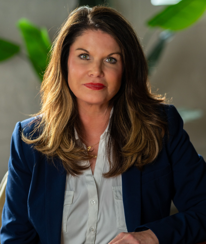A woman with long brown hair wearing a navy blazer and white blouse sits indoors. She looks directly at the camera with a slight smile, inviting you to discover Our Story. Green plants and a neutral background are visible behind her.