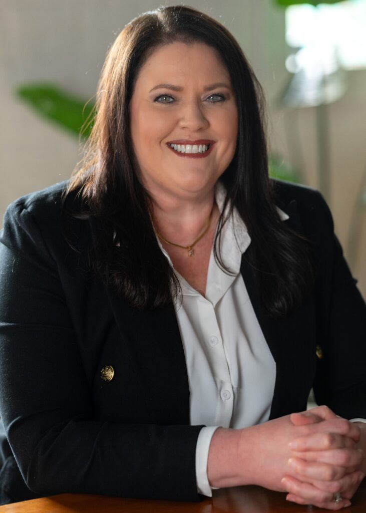 Vonda Walters, a woman with long dark hair wearing a black blazer and white blouse, sits at a table smiling with her hands clasped. The background is softly lit, with green plants visible.