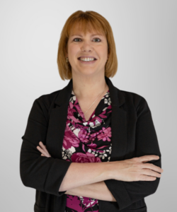 A woman with straight, light brown hair and a friendly smile stands with arms crossed. She wears a black blazer over a pink and black floral blouse, embodying confidence in revenue cycle management, against a plain, light grey background.